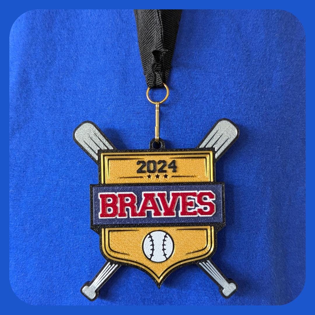 Custom 2024 baseball medal featuring a gold shield with the team name &#39;BRAVES&#39; in bold red and white letters on a black banner. The medal includes a baseball icon at the bottom and two crossed silver bats, hanging from a black ribbon